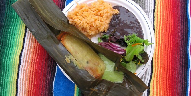 banana leaf tamale with black beans and rice on a plate, sitting on a colorful blanket