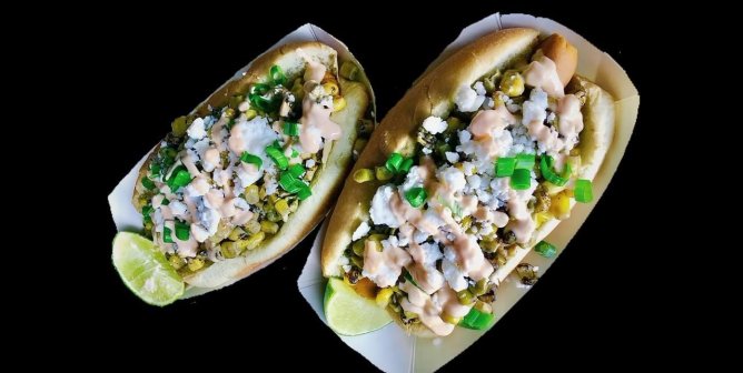 two vegan hot dogs topped with esquites, vegan feta, cilantro, and a sauce