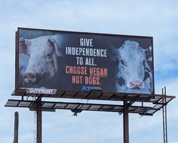Photo of billboard with images of a pig and a calf. Text reads give independence to all. Choose vegan hot dogs.