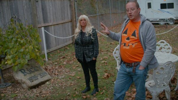 photograph. tonia haddix and cyril "cy" vierstra standing in a yard, facing viewer. Cy is gesturing to a rock headstone that reads "Ricky, 1988 to 2019"