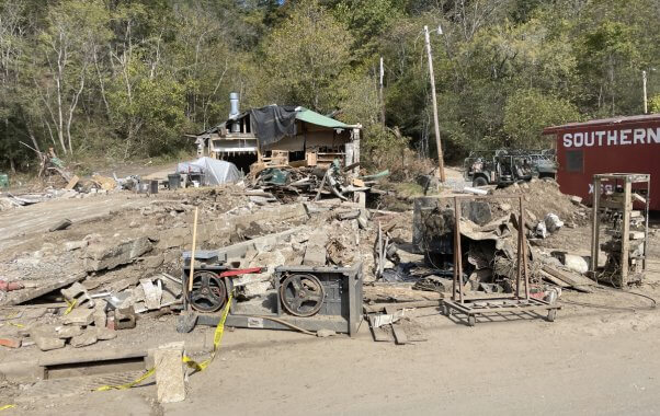 photograph of rubble in north carolina, mostly dusty brick and industrial equipment, but a half-destroyed building is visible in the background.