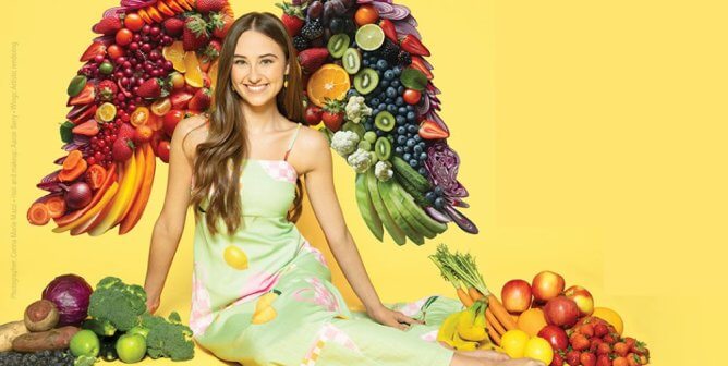 kensington tallman poses in front of vegetables/fruits shaped as angel wings