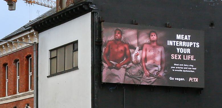 Photo of a billboard with two men in bed and a chicken between them. Text reads "Meat Interrupts Your Sex Life"